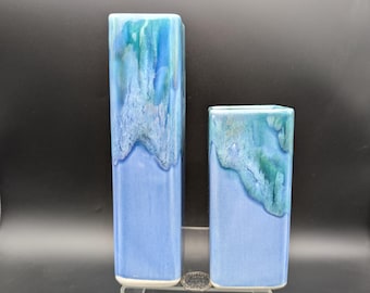 May include: Two rectangular ceramic vases. The vases are a light blue color with a darker blue and green glaze at the top, resembling a wave. The vases are displayed on a clear acrylic stand. The taller vase is approximately 10 inches tall, and the shorter vase is approximately 6 inches tall.