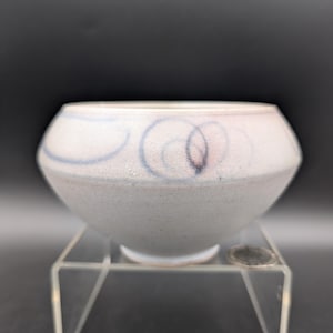 Pode incluir: Uma pequena tigela de cerâmica de cor clara, com uma abertura larga e um corpo arredondado. A tigela apresenta uma superfície texturizada e é decorada com desenhos de redemoinhos azuis e pretos. Está sobre um suporte de acrílico transparente sobre um fundo escuro.