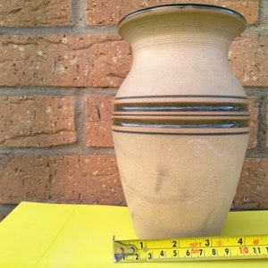 Può includere: Un vaso in ceramica beige con bordo svasato e due fasce orizzontali verde scuro attorno al centro. Un metro a nastro giallo è posizionato alla base, mostrando che il vaso ha una larghezza di circa 14 cm nel punto più largo.