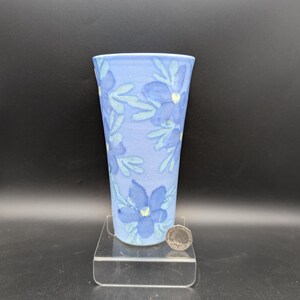 Può includere: Un vaso in ceramica conico con colore di base azzurro, decorato con un motivo ripetitivo di fiori blu scuro con centri giallo pallido e foglie azzurre. Una moneta color argento è posizionata alla base come riferimento per la scala.
