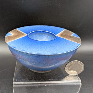 May include: A small, round, blue ceramic bowl with a wide, flat rim featuring two metallic gold-toned triangular accents. The bowl has a visible crackle glaze finish and is positioned next to a silver-colored coin for scale, showing its size.