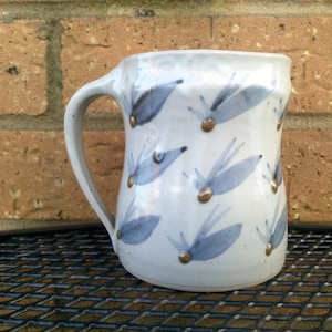 May include: A white ceramic mug with a blue and gold design. The mug has a handle and is decorated with a pattern of stylised leaves and gold dots.