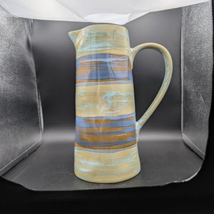 Puede incluir: Jarra de cerámica alta con asa y boquilla. La jarra presenta rayas horizontales en tonos azules, beige y marrón. Tiene un acabado liso y brillante y un asa curvada.