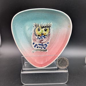 May include: A ceramic owl dish with a teal and pink gradient. The owl has green eyes, a yellow beak, and blue accents. The dish is triangular with rounded edges. It is displayed on a clear stand.