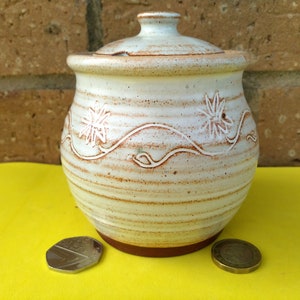 May include: A white ceramic jar with a lid, decorated with brown stripes and white floral designs. The jar is sitting on a yellow surface.