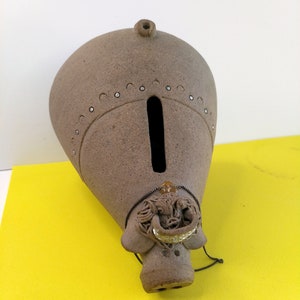 May include: A brown ceramic piggy bank with a small hole for coins. The piggy bank has a decorative design on the bottom that resembles an animal's head.