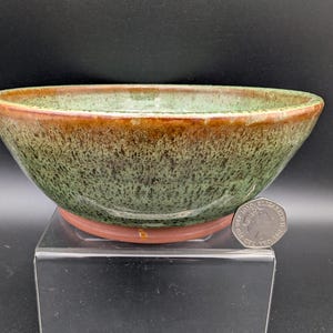 Puede incluir: Un cuenco de cerámica hecho a mano con un esmalte moteado verde y marrón. El cuenco tiene una forma ancha y poco profunda y una base marrón rojizo. Una moneda de 50 peniques está al lado del cuenco para comparar el tamaño.