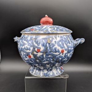 Può includere: Una ciotola da portata in ceramica con coperchio, motivo blu e bianco, accenti rossi e macchie dorate. La ciotola ha due manici e un pomello rosso sul coperchio. La ciotola è su un supporto trasparente.