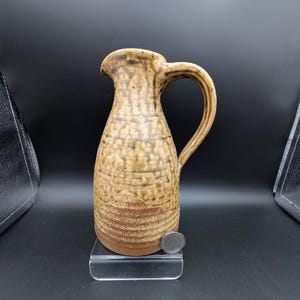 Puede incluir: Jarra de cerámica hecha a mano con superficie texturizada y asa. La jarra presenta una combinación de colores de dos tonos, con una parte superior marrón claro y una base marrón más oscura. La jarra mide aproximadamente 20 cm de altura.