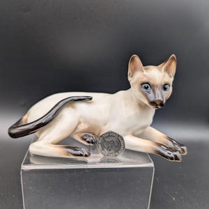 Op de afbeelding: Een keramieken Siamese kattenfiguur in een liggende houding. De kat heeft een crèmekleurig lichaam met bruine aftekeningen op het gezicht, oren, poten en staart. Het heeft blauwe ogen en rust op een heldere, rechthoekige standaard.
