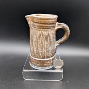 May include: A small, textured ceramic pitcher in shades of brown and tan, with a handle and a spout. The pitcher has a woven pattern on the body and a light blue accent at the base. A coin is next to the pitcher for size reference.