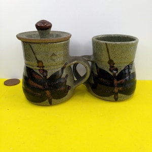 May include: A ceramic sugar bowl and creamer set. The set is in a muted green with brown dragonfly designs. The sugar bowl has a lid with a small brown knob. The creamer and sugar bowl are connected by a handle.