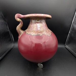 May include: A round ceramic pitcher with a glossy deep red base and a textured, mottled brown and tan glaze around the neck and rim. The vessel features a single curved handle on the left side and a wide opening. A small silver coin sits at the base.