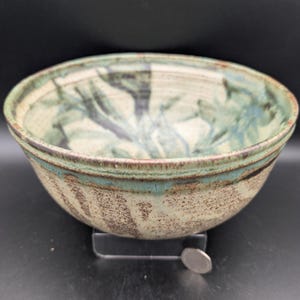 Könnte beinhalten: Eine handgefertigte Keramikschale mit breitem Rand und strukturierter Oberfläche. Die Schale hat eine cremefarbene Basis mit blau-grünen blattartigen Mustern im Inneren. Der Rand hat eine türkisfarbene Glasur. Eine Münze liegt neben der Schale.