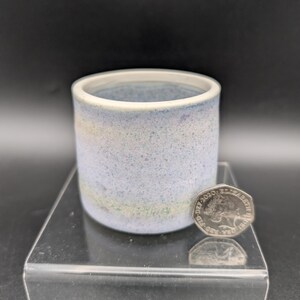 May include: A small, cylindrical ceramic cup with a speckled blue and white glaze. The cup has a white rim and a light blue interior. A coin is next to the cup for size comparison.
