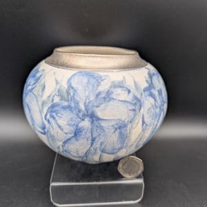 May include: A round ceramic vase with a silver rim and a white base. The vase is decorated with blue floral patterns. A coin rests on a clear stand in front of the vase.