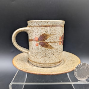 Op de afbeelding: Een gespikkelde keramische koffiekop en schotel set. De beker heeft een bruin en oranje bladeren ontwerp. De ronde schotel past bij de gespikkelde afwerking van de beker. De set staat op een transparante displaystandaard.