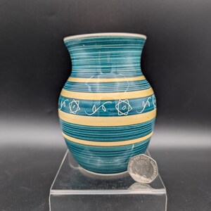 May include: A small, turquoise and yellow ceramic vase with a rounded body and a narrow neck. The vase features horizontal stripes and decorative star-like patterns. A coin rests on the clear display stand.