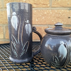 Peut inclure: Un ensemble de deux tasses en céramique avec un motif floral. La tasse de gauche a une anse et est plus haute que la tasse de droite. Les deux tasses sont décorées de fleurs blanches et de feuilles vertes sur un fond gris foncé.