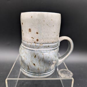 May include: A handcrafted ceramic mug with a distinctive two-tone appearance. The upper section is a light, speckled off-white, and the lower section is a textured blue-grey. It features a curved handle and is displayed on a clear acrylic stand.