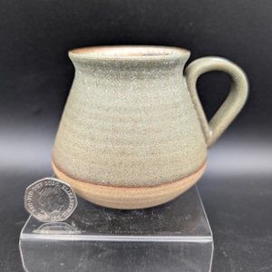 Può includere: Una tazza in ceramica con una smaltatura testurizzata color verde salvia sulla parte superiore e una base liscia color terracotta non smaltata. La tazza presenta un manico curvo ed è posizionata accanto a una moneta britannica da 50 pence del 2020.