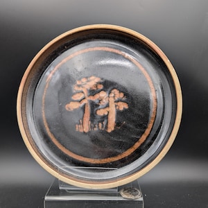 May include: A round, ceramic plate with a dark brown center and a lighter brown rim. The center features a design of two stylized trees in a lighter brown color. The plate is displayed on a clear stand.