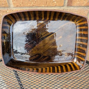 May include: Rectangular ceramic serving dish with a glossy, dark brown interior and a striped pattern in shades of brown and orange around the edges. The dish is placed on a metal grate.