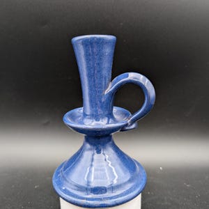 May include: A blue ceramic candle holder with a handle. The holder has a wide base and a narrow top with a small opening for a candle.