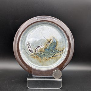 May include: A round ceramic plate with a dark brown rim and a speckled light blue and grey centre. The plate features a hand-painted design of dark blue wheat stalks and stylised leaves. A silver coin is placed next to the plate for scale.