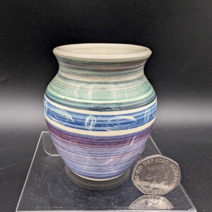 May include: A small, hand-painted ceramic vase with a rounded body and a slightly flared rim. The vase features horizontal stripes in shades of blue, green, and purple, with white decorative lines. A coin is next to the vase.