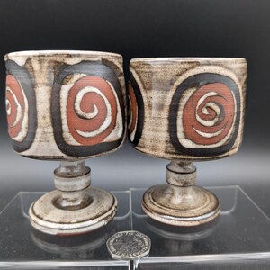 May include: Two ceramic goblets with a spiral design in red, black, and white. The goblets have a cylindrical bowl and a pedestal base. The background is black.