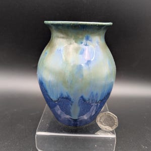 Könnte beinhalten: Eine kleine, handgefertigte Keramikvase mit rundem Körper und ausgestelltem Rand. Die Vase hat eine Mischung aus blauer, grüner und weißer Glasur, die einen einzigartigen Tropfeffekt erzeugt. Die Vase ist etwa 10 cm hoch.