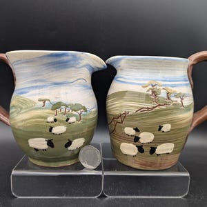 May include: Two ceramic jugs with brown handles. Each jug has a painted landscape with sheep grazing on a green hillside under a blue and white sky. The jugs are displayed on a clear stand.