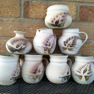 Könnte beinhalten: Eine Sammlung von acht Keramikbechern und einer kleinen Vase, alle mit einer weißen Glasur und handbemalten braunen und rosafarbenen Blumendesigns.