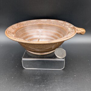 May include: A handmade, brown ceramic bowl with a handle. The bowl has a circular shape with a decorative rim. The bowl is approximately 6 inches in diameter and 2 inches deep. The bowl is sitting on a clear acrylic stand.