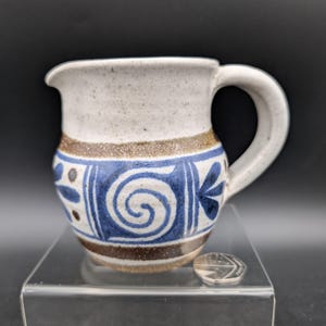 May include: A small, handmade ceramic pitcher with a white speckled top and a blue and brown patterned design. The pitcher features a handle and a spout for pouring. The design includes a blue spiral and floral motifs.