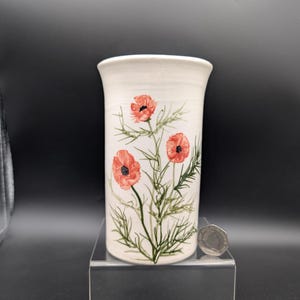 May include: A white ceramic vase with a flared top, adorned with hand-painted red poppy flowers and green foliage. The vase is approximately 6 inches tall and has a cylindrical shape. The design is simple and elegant, suitable for displaying flowers.