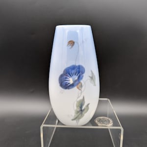 May include: A white and blue vase with a floral design. The vase features a blue flower with green leaves and brown stems. The vase is on a clear acrylic stand.