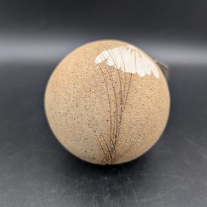 Peut inclure: Objet rond en céramique beige avec un motif floral blanc et des accents linéaires noirs. L'objet a une surface texturée et semble être une pièce décorative ou un petit récipient. Le fond est gris foncé.