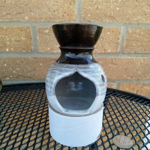 May include: A brown and white ceramic oil burner with a round body and a wide opening at the top. The burner has a cutout in the front that resembles a teardrop shape. The burner is sitting on a white cardboard base.