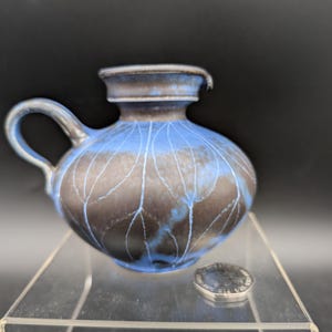 May include: A small ceramic jug with a rounded body and a handle. The jug is primarily blue with brown accents and a leaf-like pattern. A small, silver-coloured lid sits nearby. The jug is displayed on a clear surface.