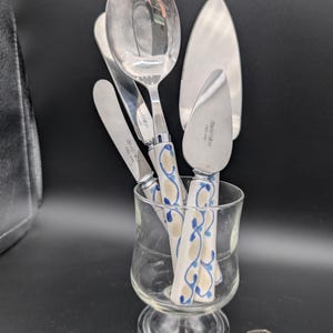 May include: Silver cutlery set including a spoon, knives, and a cake server, presented in a clear glass. The knife and spoon handles have a white base with a blue and gold floral pattern. The items are set against a black background.