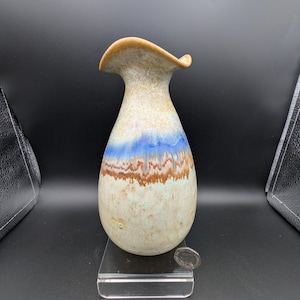 Könnte beinhalten: Eine Keramikvase mit ausgestelltem Rand und bauchigem Körper. Die Vase hat eine Farbpalette aus Beige, Blau und Braun. Der Rand ist in einem warmen Beigeton gehalten. Die Vase ist etwa 18 cm hoch.