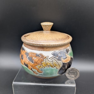 May include: A ceramic jar with a lid, featuring a brown speckled finish. The jar's body showcases a hand-painted design of two pigs in brown, black, and white, set against a green and white background. The lid has a small knob.