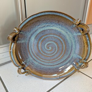 May include: A handmade ceramic serving platter with a spiral design in shades of brown and blue. The rim and handles are a darker brown, and the centre features a lighter blue swirl. The platter has four decorative handles.