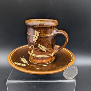 May include: A brown ceramic teacup and saucer set. The cup and saucer are decorated with wheat stalk designs in a lighter brown color. The set is on a black surface.