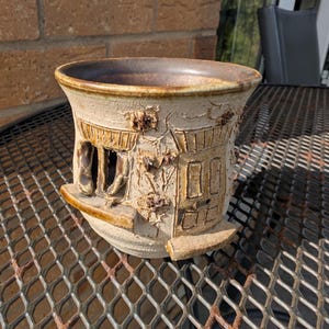 May include: A beige ceramic planter with a textured surface featuring relief carvings of a house facade, including a window and a door. The rim is dark brown, and the exterior has a rough, matte finish with small raised details resembling foliage.