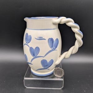 Op de afbeelding: Een keramische kan met een witte basis en een blauw bloemmotief. De handgreep heeft een gedraaid touwontwerp. De kan staat op een transparante acrylstandaard.