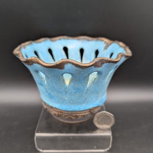 May include: A ceramic bowl with a bright blue glazed exterior and a dark brown rim. The bowl features a scalloped edge and a series of vertical cutouts around the upper section. A small silver coin is placed next to the base for scale.