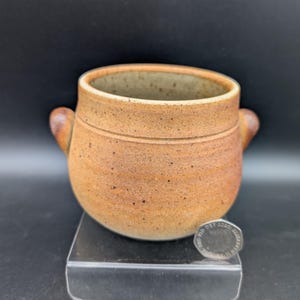 May include: A small, round, speckled tan ceramic pot with two side handles and a wide opening. The exterior features a textured, earthy finish with subtle horizontal ridges. A British twenty pence coin is placed next to the pot for scale.